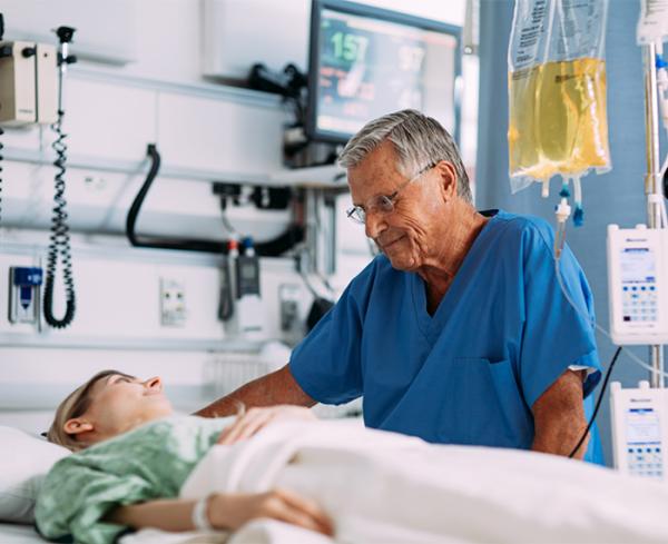 A healthcare professional interacts with a patient at the bedside. An IV bag and infusion pumps are shown in the background.