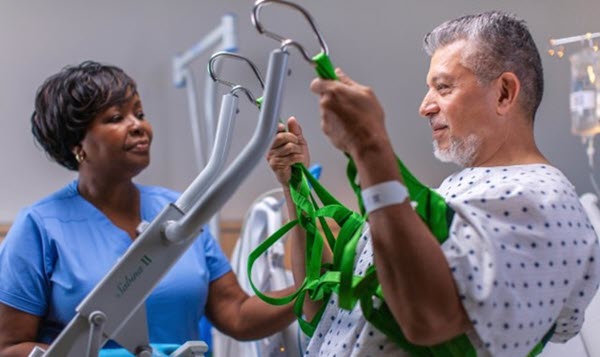 A male patient uses a mobility lift with assistance from a healthcare professional