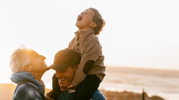 A couple and their grandson, who is on his grandfather's shoulders, laughing at the beach
