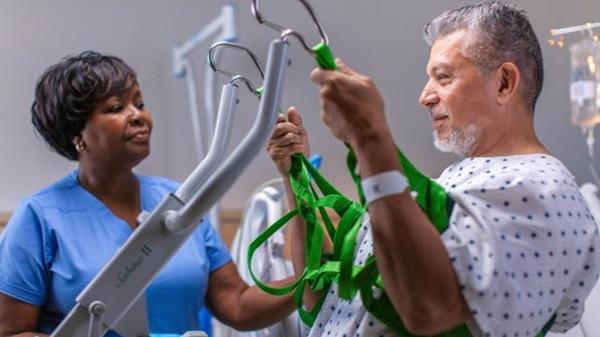 A male patient uses a mobility lift with assistance from a healthcare professional