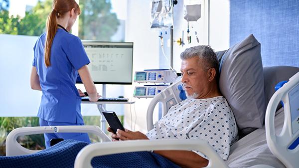 A patient in a Baxter hospital bed uses a tablet while a clinician works at a nearby computer.