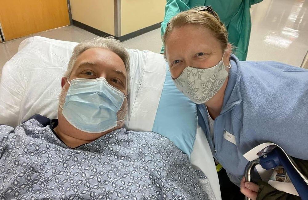 Baxter employee masked and in hospital bed with wife by his side