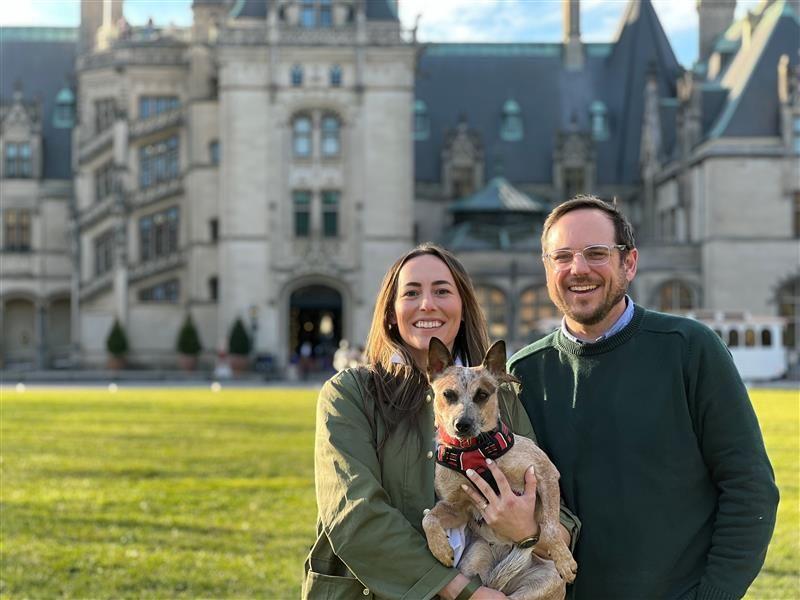 A couple holding a dog stands in front of a palatial estate