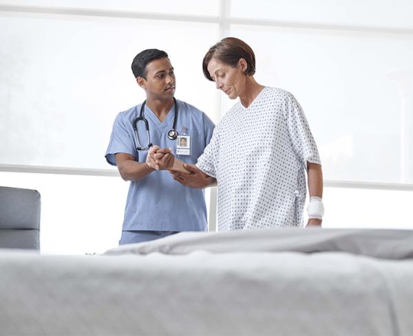 A doctor helps their patient in a hospital gown take a few steps around their hospital room