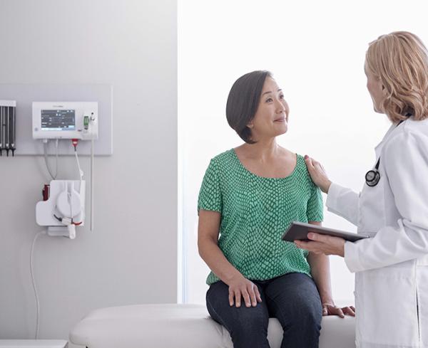 A patient sits on an exam table in a doctor's office. The doctor has a hand placed on the patient's shoulder