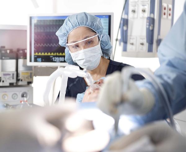 An anesthesiologist sits behind the patient in the operating room while we see two pairs of gloved hands operating on the patient in the foreground 