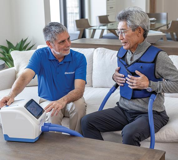 an adult patient wears The Vest APX System vest garment at home, sitting next to a Baxter representative