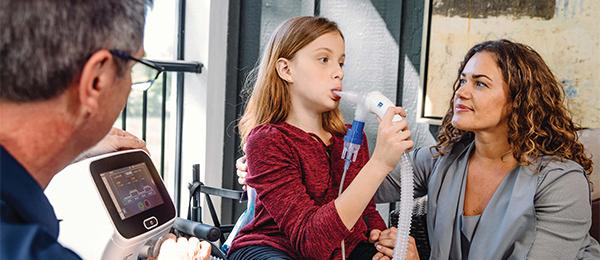A young girl uses a respirator care device in a doctor's office while her mother and a healthcare professional watch encouragingly