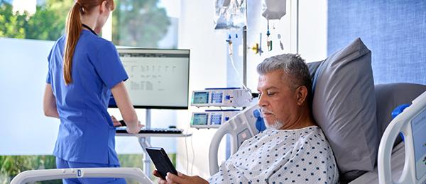 A patient in a Baxter hospital bed uses a tablet while a clinician works at a nearby computer.